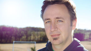 Close-up portrait of a man with light eyes and a small goatee, smiling slightly against a sunny outdoor background.