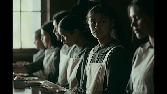 Several young women wearing white aprons stand together in a dark, indoor setting.