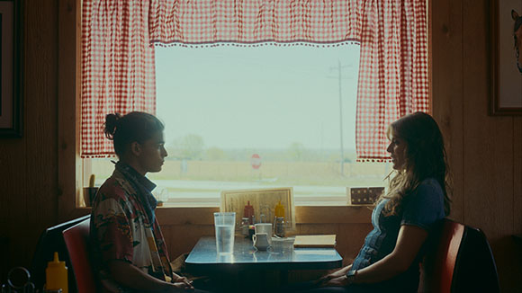 Two women sit facing each other at a small table in front of a large window with red checkered curtains.