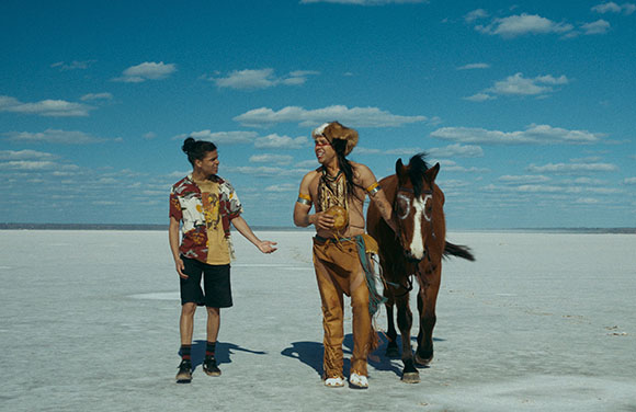 Two men walk across a vast white salt flat under a blue sky with a brown horse walking beside them.