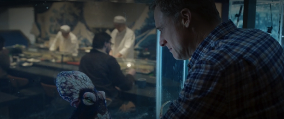 A man in a plaid shirt looks through a glass tank at a restaurant with chefs in the background.