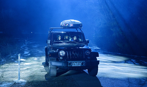 A dark SUV with a roof cargo box drives down a road illuminated by bright blue light in a dark forest.
