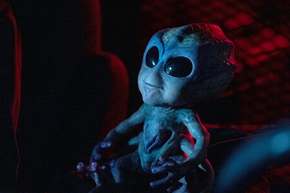 A small, blue-skinned alien creature with large black eyes sitting in dark red lighting.