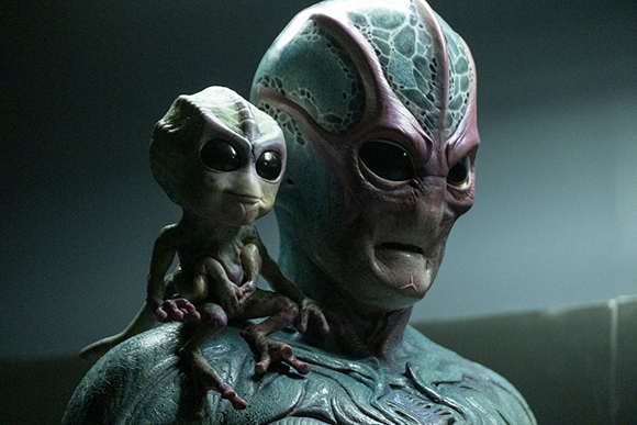 A small, pale alien sits on the shoulder of a larger, textured alien with dark eyes.