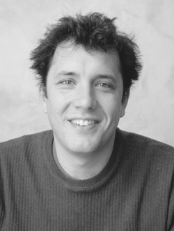 Black and white portrait of a smiling man with dark, wavy hair wearing a textured sweater.