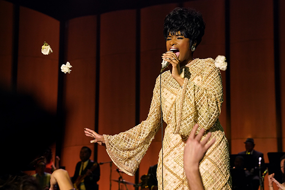 A woman in a shimmering gold gown sings into a microphone as white flowers fall around her on a dark stage.