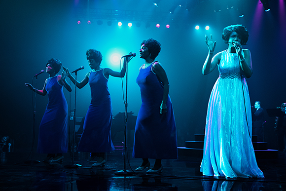 Four women in long dresses sing into microphones on a stage under blue and teal spotlights.