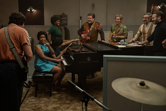 Musicians in a recording studio laughing and playing instruments around a black grand piano.