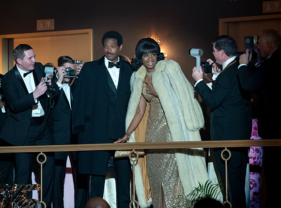 A woman in a gold gown and white fur coat smiles while being photographed by several men in tuxedos.