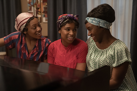 Three Black women in colorful headwraps and patterned dresses smiling while looking at a piano.