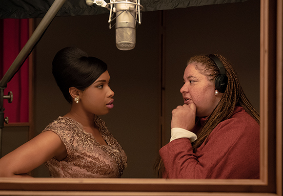 A woman in a sequined dress looks at a woman wearing headphones in a recording studio.