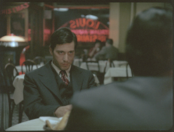 A man in a dark suit and tie sits at a restaurant table, looking toward a person in the foreground.