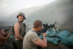 Soldiers in tactical gear aiming a machine gun from a fortified position overlooking a mountain valley.