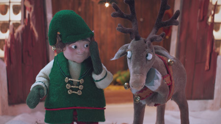 A felt elf in green clothing looks at a felt reindeer in a snowy setting.