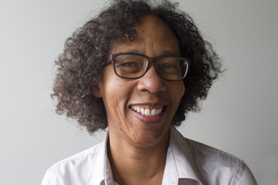 A smiling woman with dark curly hair and glasses wearing a light-colored collared shirt.