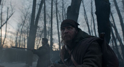 A bearded man wearing a dark headscarf and heavy coat looks intensely into the distance in a dim forest.