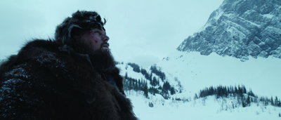 A man with a thick beard and fur clothing looks out over a snowy mountain landscape.