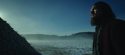 A bearded man in a dark coat looks across a misty, snow-covered field toward a distant treeline under a blue sky.