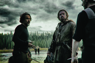 Three men in rugged clothing stand in a field under a dark, cloudy sky near a forest.