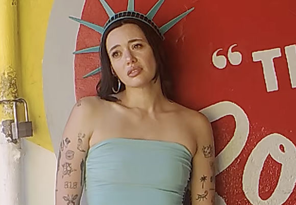 A woman in a light blue tube top and Statue of Liberty crown leans against a red wall with white text.