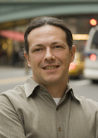 A smiling man with long dark hair pulled back, wearing a tan button-down shirt in a city setting.