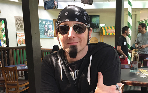 A man with a black bandana, sunglasses, and a goatee smiles while sitting at an outdoor restaurant table.
