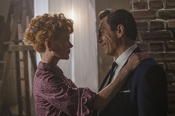 A woman with curly red hair touches the shoulder of a man in a suit as they gaze at one another.