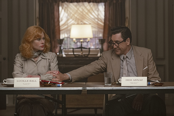 A man in a suit and a woman with red hair sit at a table with nameplates reading Lucille Ball and Desi Arnaz.