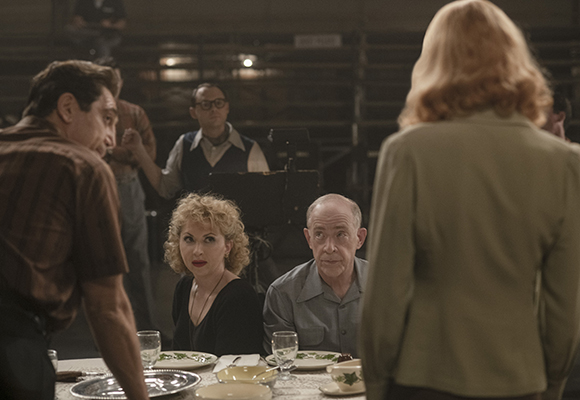 A man in a brown shirt leans toward a woman with blonde hair at a dining table in a dark room.