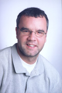 A man with short dark hair and glasses wearing a light grey polo shirt against a white background.
