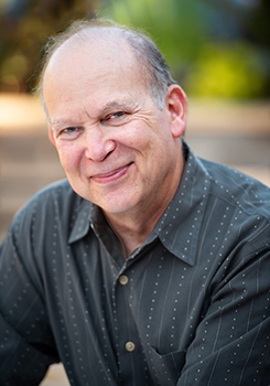 A smiling man with light skin and thinning hair wearing a dark patterned button-down shirt.