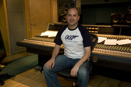 A man in a 007 t-shirt sits in a chair before a large professional audio mixing console.