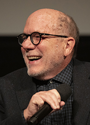 A bald, smiling man with glasses and a grey beard holds a microphone near his mouth.