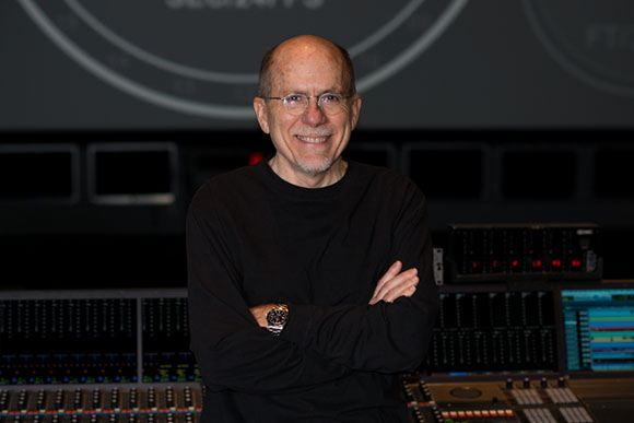 A man with glasses and a graying beard smiling with arms crossed in a studio with mixing consoles.