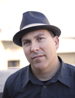 A man in a black fedora and black shirt looking toward the camera.