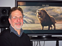 A man with glasses and a beard smiles beside a large screen showing a roaring lion in a landscape.
