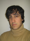 A man with dark, wavy hair wearing a tan turtleneck sweater against a plain background.