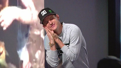 A man wearing a black DC cap and grey sweatshirt leans forward with his hands pressed against his face.
