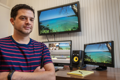 A man in a red and blue striped shirt stands in a room with three monitors displaying tropical scenery.