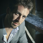 Close-up portrait of a man with short dark hair and facial hair wearing a grey suit jacket and white shirt.