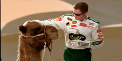 A man in a white racing suit pets a camel's head in a sandy desert landscape.