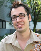 A smiling man with dark spiked hair and glasses wearing a tan patterned shirt.