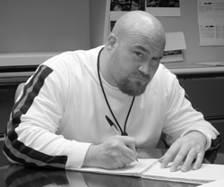 A bald man with a goatee wearing a long-sleeved shirt writes in a notebook at a desk.