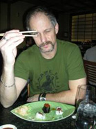 A man in a green t-shirt holds chopsticks up to his eye while sitting at a table with food.