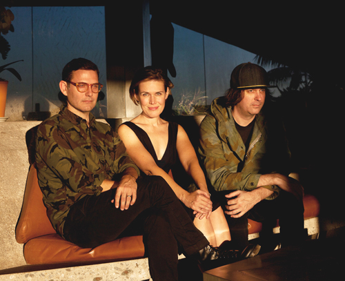 A man in a camo shirt, a woman in a black tank top, and a man in a green jacket sit on a tan sofa.