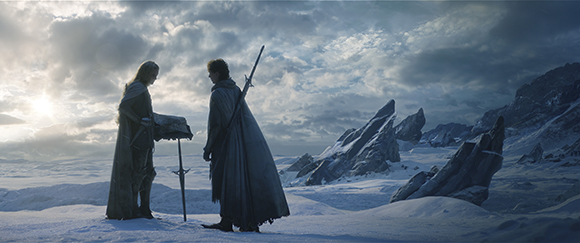 Two cloaked figures stand in a vast, snowy mountain landscape under a cloudy sky.