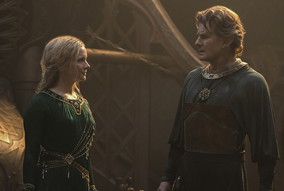 A blonde woman in a green velvet gown looks at a man in dark green and brown period clothing.