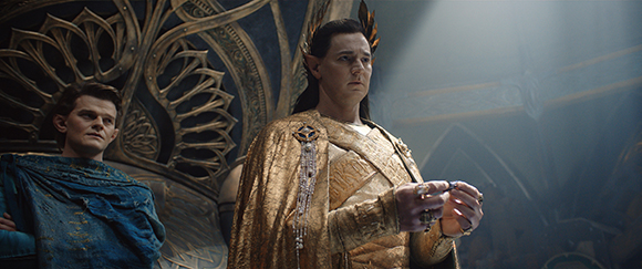 A man with pointed ears in gold armor stands before another man in blue robes in a dimly lit hall.