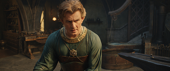 A man with blond hair wearing a teal and gold tunic looks forward with a serious expression.