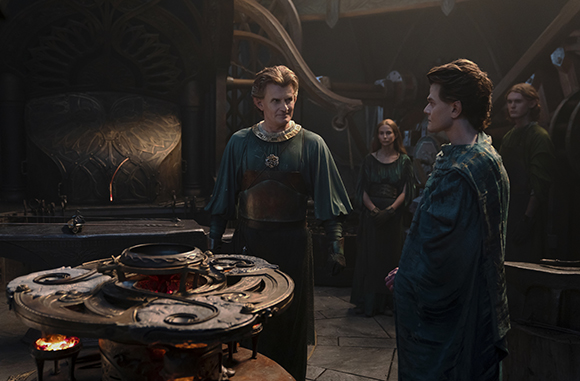 Four people in period costumes stand in a dark, intricately decorated room with large metal structures.